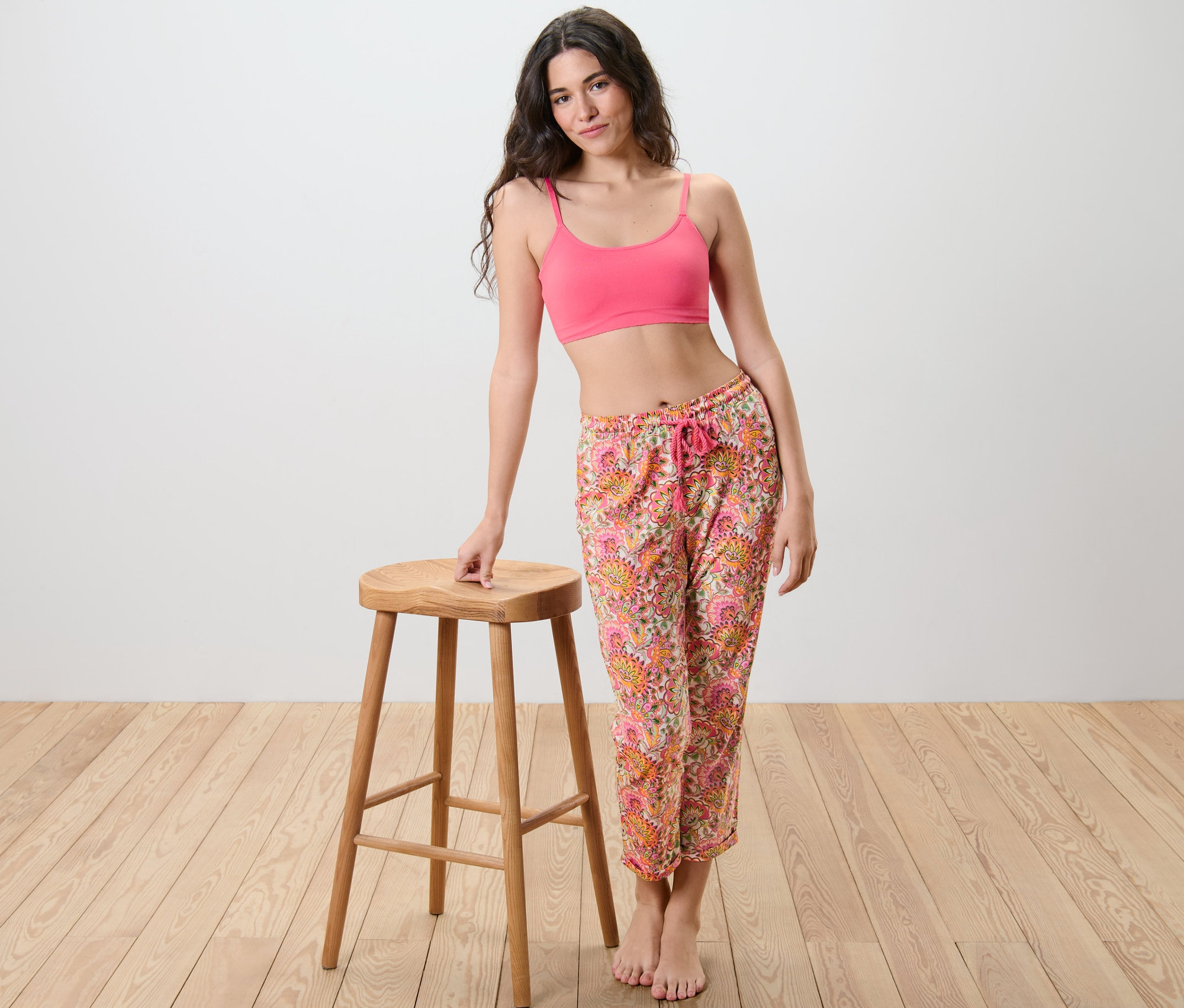 Frau posiert neben einem Hocker, trägt ein pinkes Top und eine Pyjamahose.