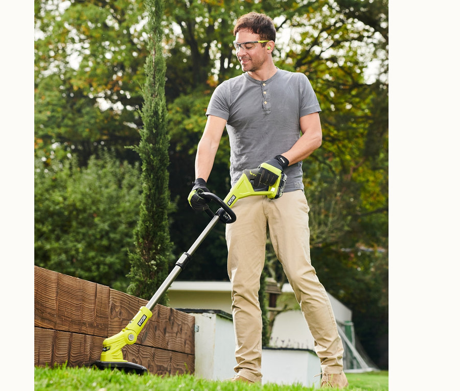Ein Mann mit Schutzbrille und Handschuhen trimmt mit einem Akku-Rasentrimmer den Rasen entlang einer Holzbordüre. Im Fokus steht ein RYOBI 18 V One+ Akku-City-Rasenmäher-Set »RY18LMC30A-120«.