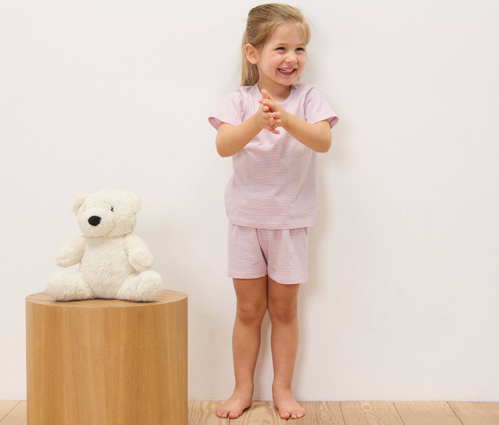 Lächelndes Mädchen klatscht in kurzem Kinder-Pyjama (2er-Pack) mit Streifen. Links sitzt ein Teddybär auf einem Holzhocker.