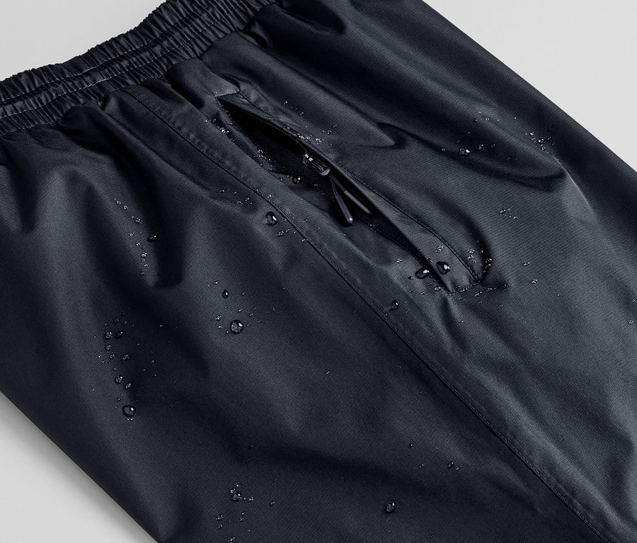 Detailaufnahme einer schwarzen Unisex-Regenhose mit Wassertropfen und Reissverschlusstasche.