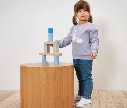 Ein kleines Mädchen spielt mit Holzbausteinen auf einem runden Tisch. Sie trägt eine Kinder-Jeans – Fit »Lotta«.