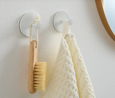 An einer weißen Wand hängen zwei Haken. An einem hängt eine Holzbürste und am anderen ein weißes Handtuch.