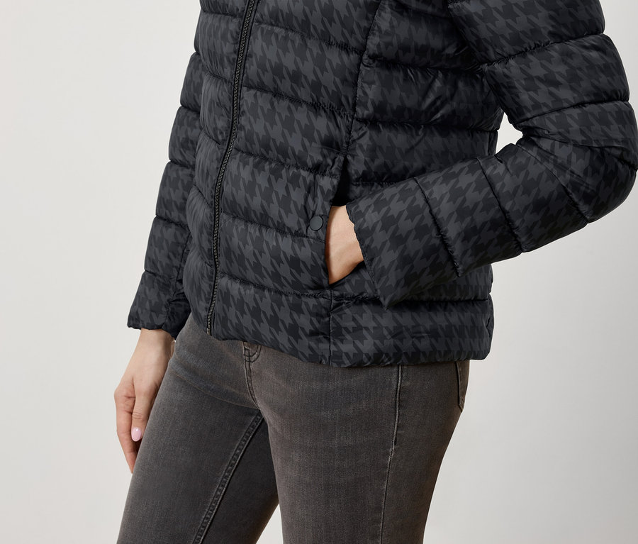 Frau hat die Hand in der Tasche einer schwarzen Daunenjacke mit Hahnentrittmuster und Jeans.