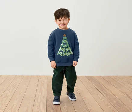 Ein lächelnder Junge steht in einem blauen Pullover mit einem Weihnachtsbaum aus Gamecontrollern, grünen Hosen und blauen Schuhen.