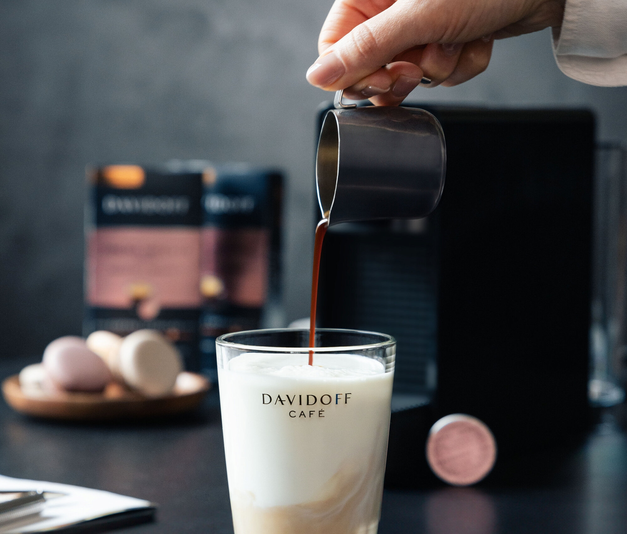 Eine Hand gießt Kaffee in ein Glas Milch. Im Hintergrund befinden sich eine Kaffeemaschine und eine Packung DAVIDOFF CAFÉ Crema Intense.