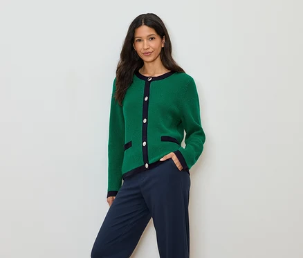 Eine Frau mit dunklen Haaren trägt eine grüne Strickjacke mit marineblauen Akzenten und marineblaue Hosen.
