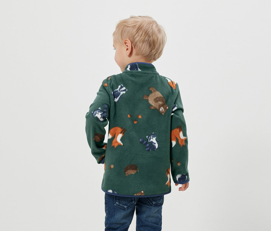 Ein Junge mit dem Rücken zur Kamera trägt eine grüne Fleecejacke mit Tiermuster und Jeans.