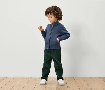 Junge mit lockigem Haar steht und hält einen kleinen Gegenstand in der Hand. Er trägt einen blauen Strickpullover mit Reißverschluss, grüne Cordhosen und weiße Turnschuhe.
