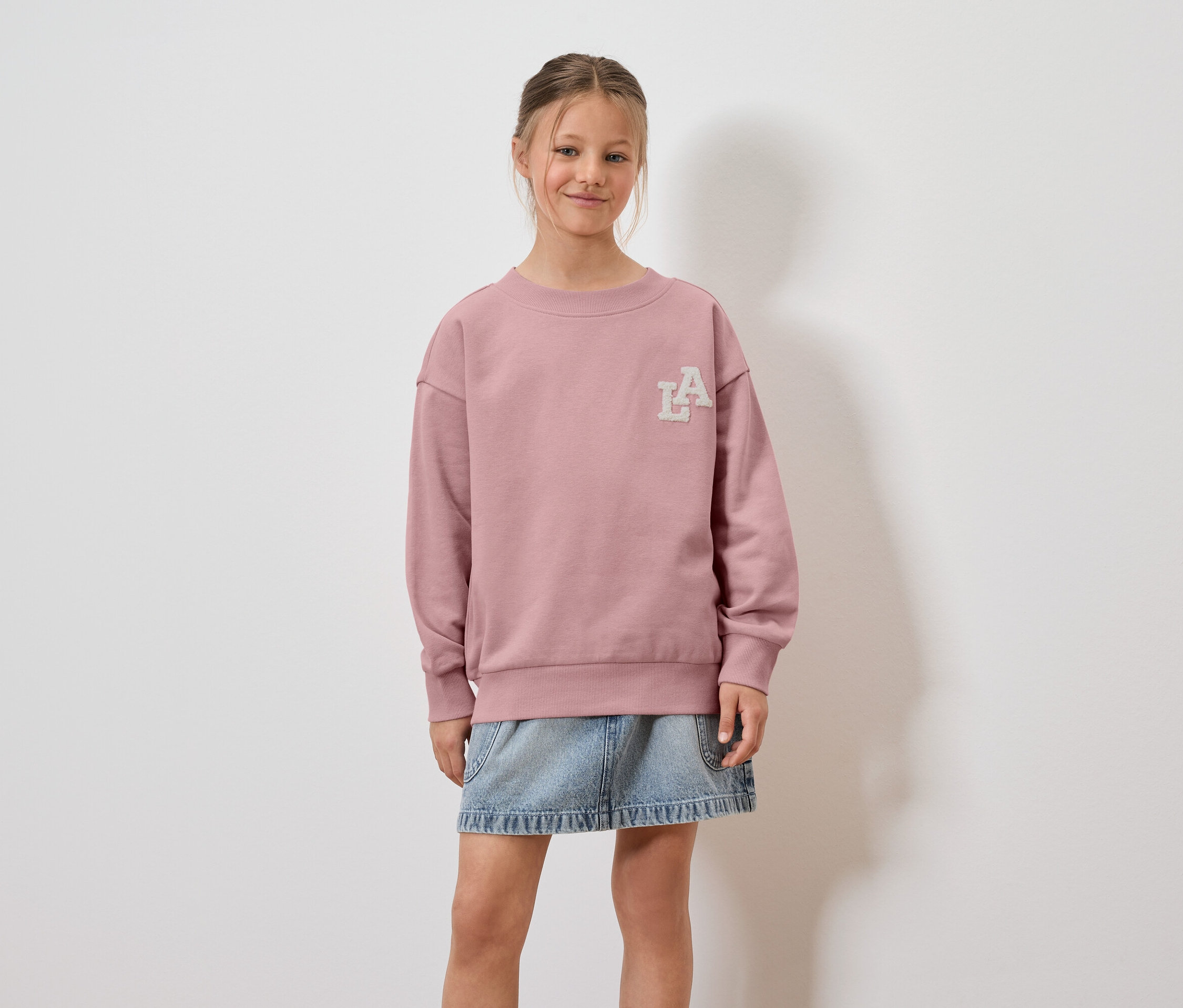 Mädchen trägt rosa Sweatshirt mit LA-Aufdruck und Jeansrock.