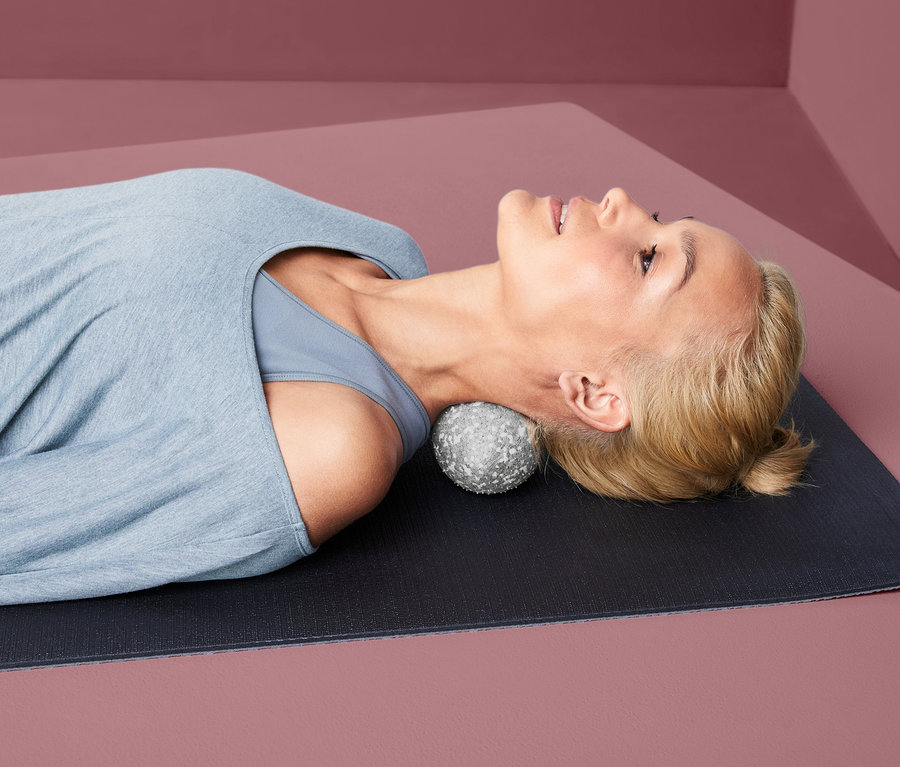 Frau liegt auf einer Matte, den Kopf auf einem Duo-Faszienmassageball.