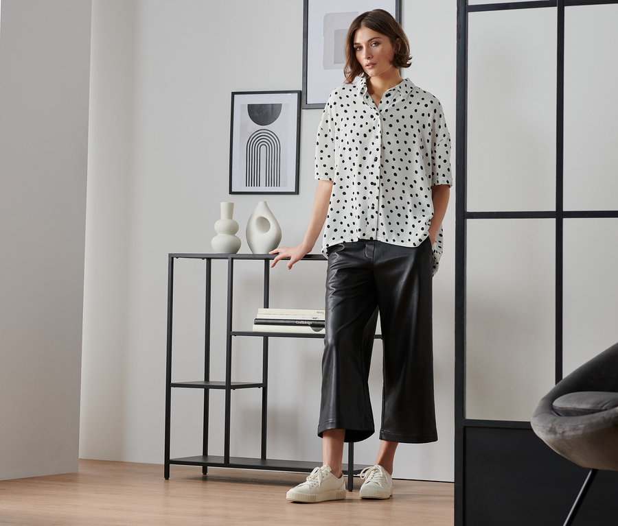 Frau steht im Raum und trägt eine kurzärmlige Webbluse mit schwarzen Punkten.