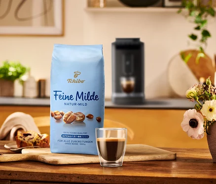 Eine Packung Tchibo Feine Milde Kaffee steht neben einer Tasse Espresso und einem Stück Kuchen auf einem Holztisch.
