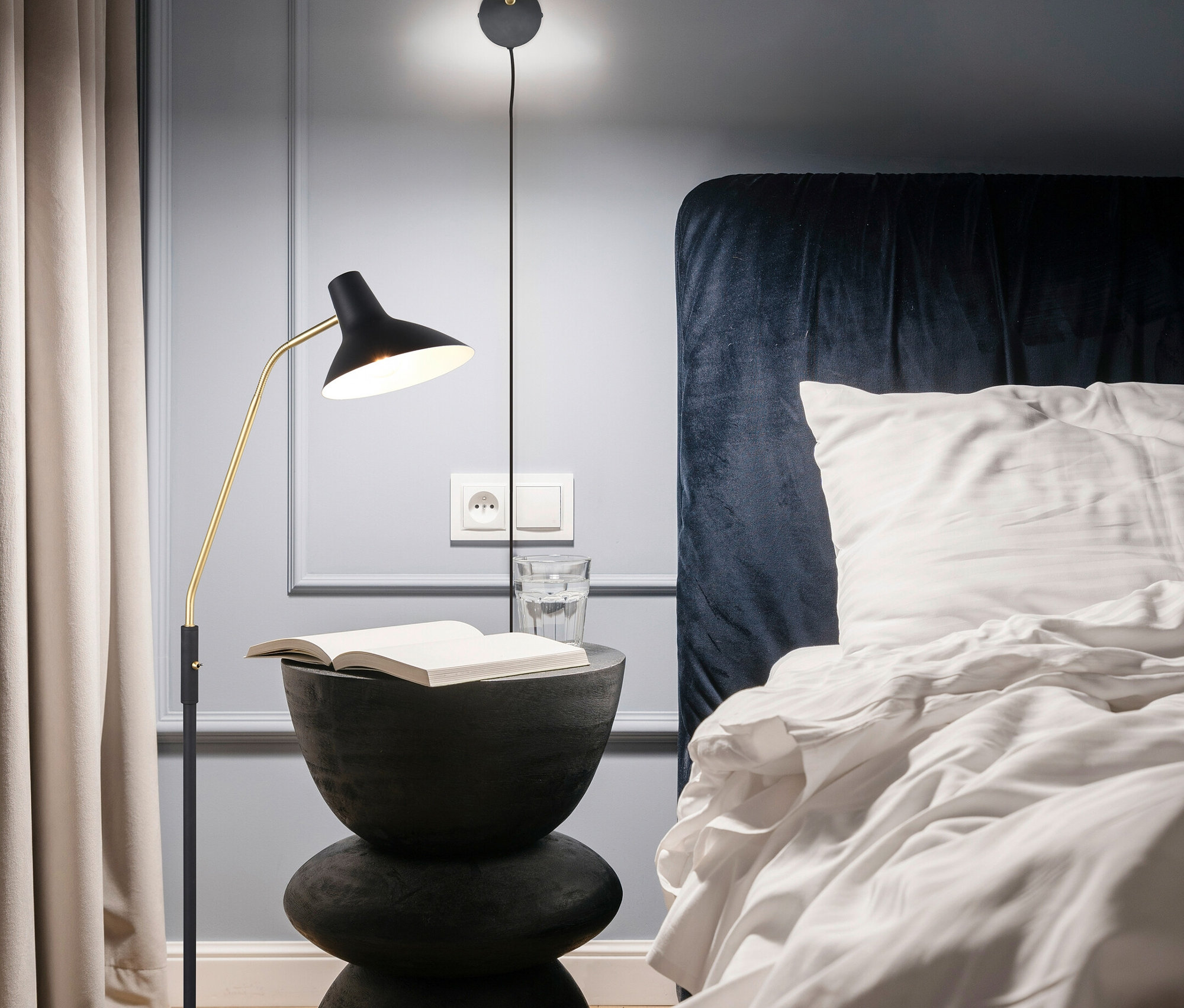 Nachttisch mit Buch und Wasserglas neben einem Bett mit blauem Kopfteil. An der Wand hängt eine Wandlampe.