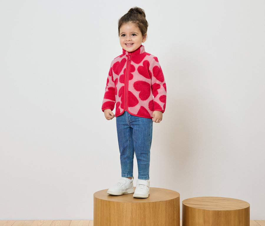 Lächelndes kleines Mädchen steht auf einem Holzsockel und trägt eine rosa Fleecejacke mit rotem Herzmuster, Jeans und weiße Turnschuhe.