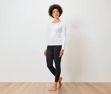 Eine Frau steht und trägt ein weißes Langarmshirt und schwarze Leggings.