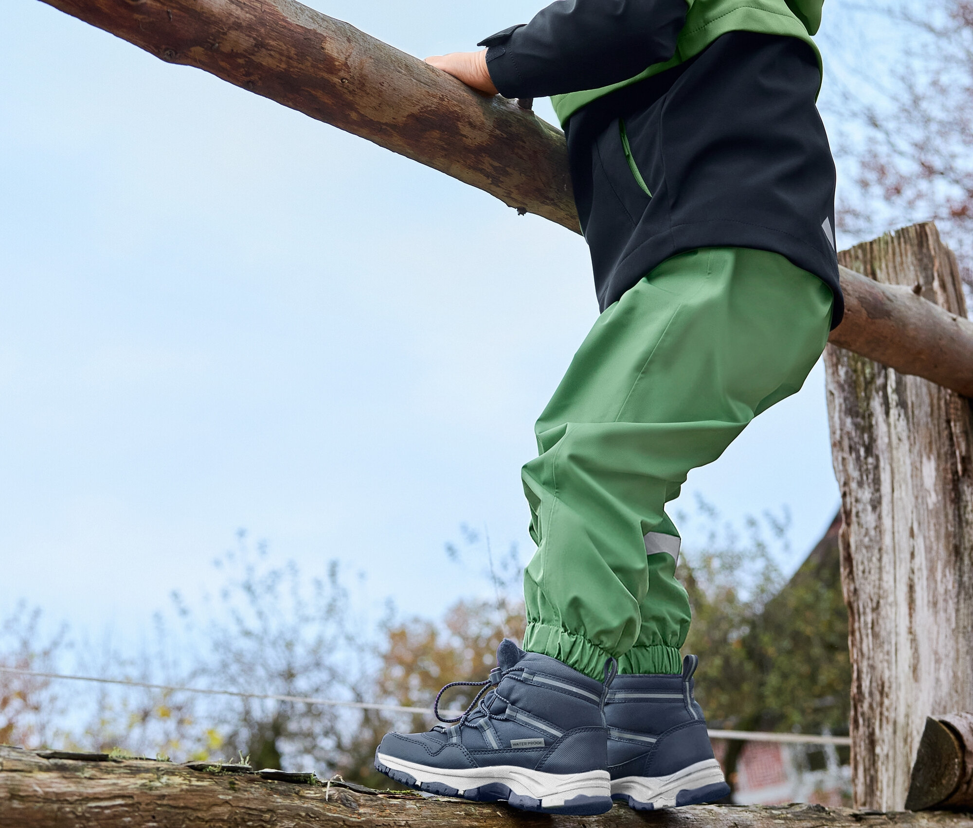 Ein Junge klettert auf einen Holzzaun. Er trägt eine grasgrüne Kinder-Regenhose, grüne Kinder-Thermostiefel und eine Kinder-Softshelljacke.