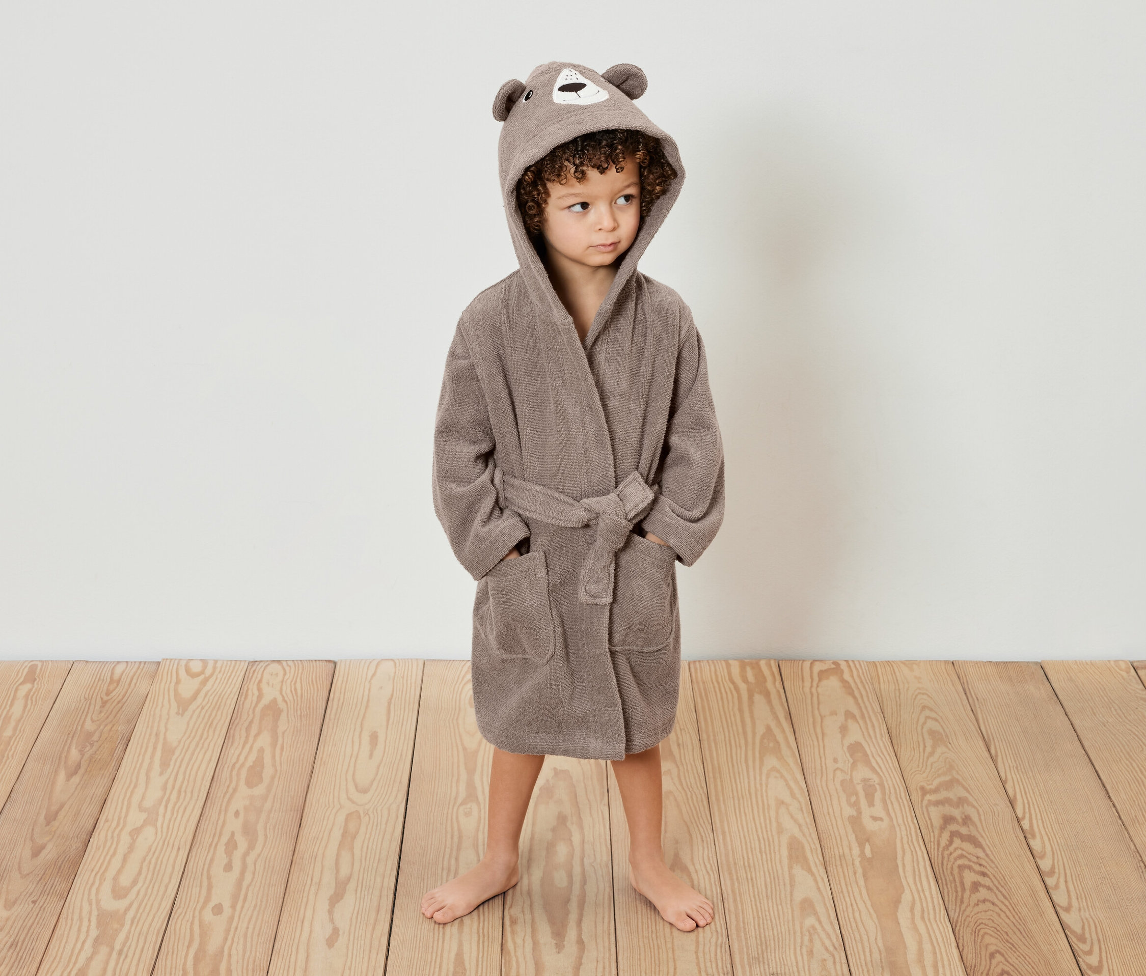 Ein Kind mit lockigem Haar steht auf einem Holzboden und trägt einen braunen Bademantel mit Bärenohren-Kapuze.