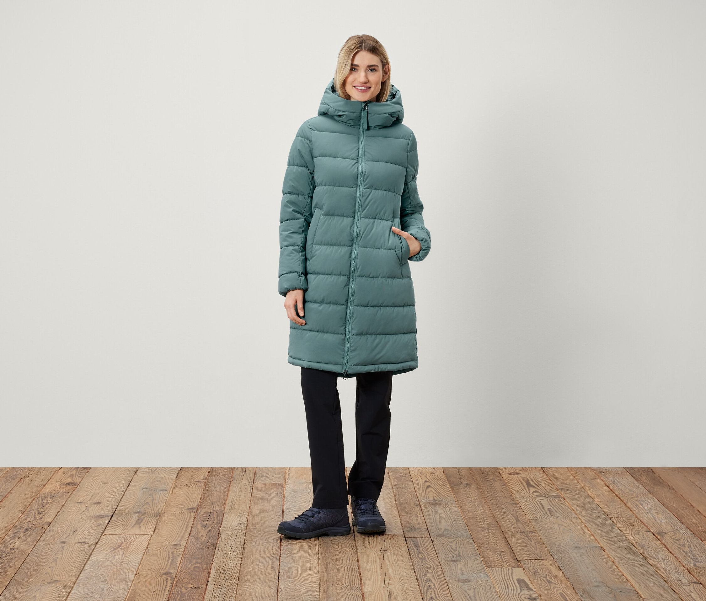 Eine Frau steht und trägt einen langen Steppmantel mit Kapuze, eine schwarze Hose und Schuhe.