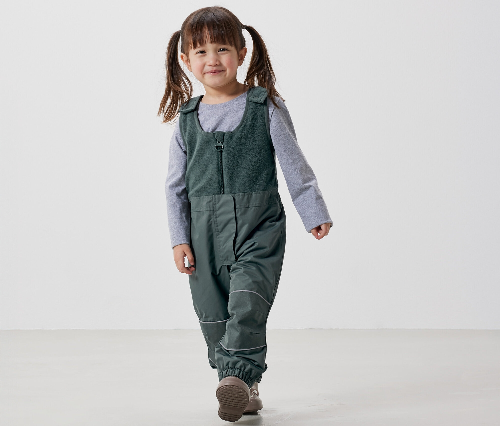 Lächelndes Mädchen mit Zöpfen geht in grüner Latzhose und grauem T-Shirt vor einem weißen Hintergrund.