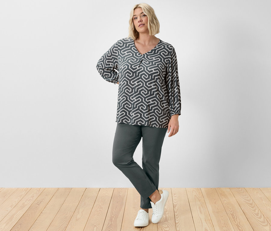 Eine Frau posiert im Studio in dunkelgrüner Stretchhose und Bluse in Knitteroptik.