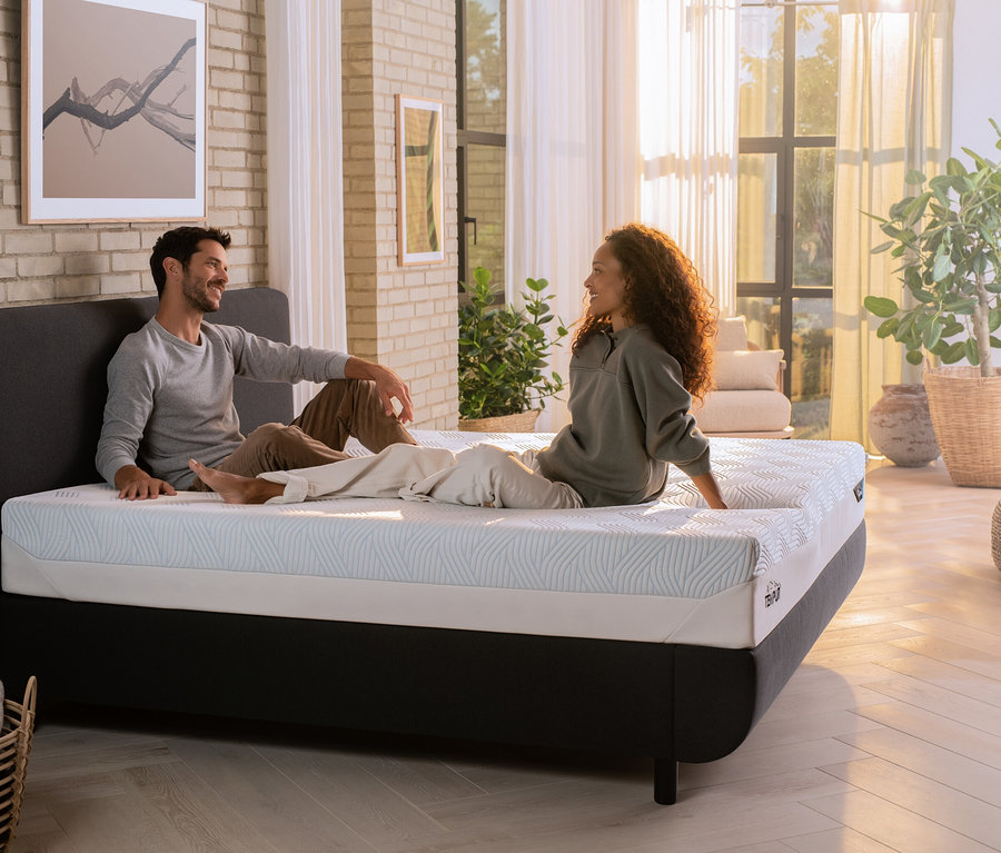 Ein Mann und eine Frau sitzen auf einer Tempur Matratze Prima in einem Schlafzimmer.