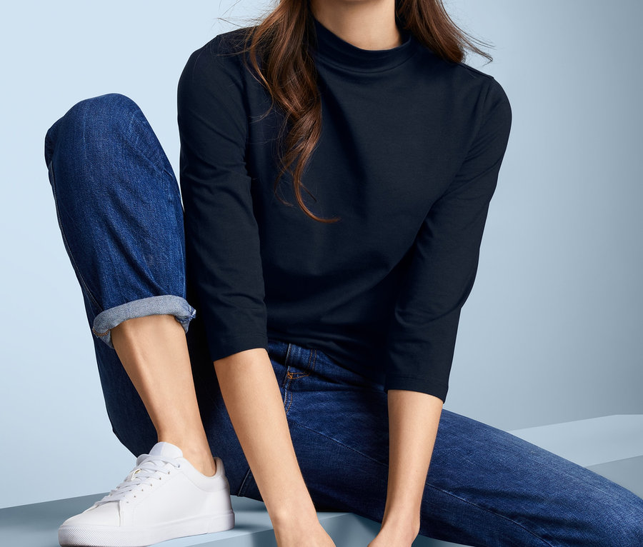 Frau sitzt und trägt ein Shirt mit Stehkragen in Dunkelblau, Jeans und weiße Turnschuhe.