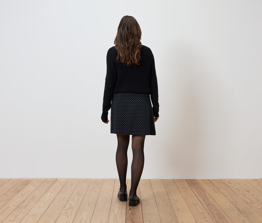 Eine Frau steht mit dem Rücken zur Kamera, trägt einen schwarzen Pullover, einen gemusterten Rock und eine schwarze Strumpfhose.