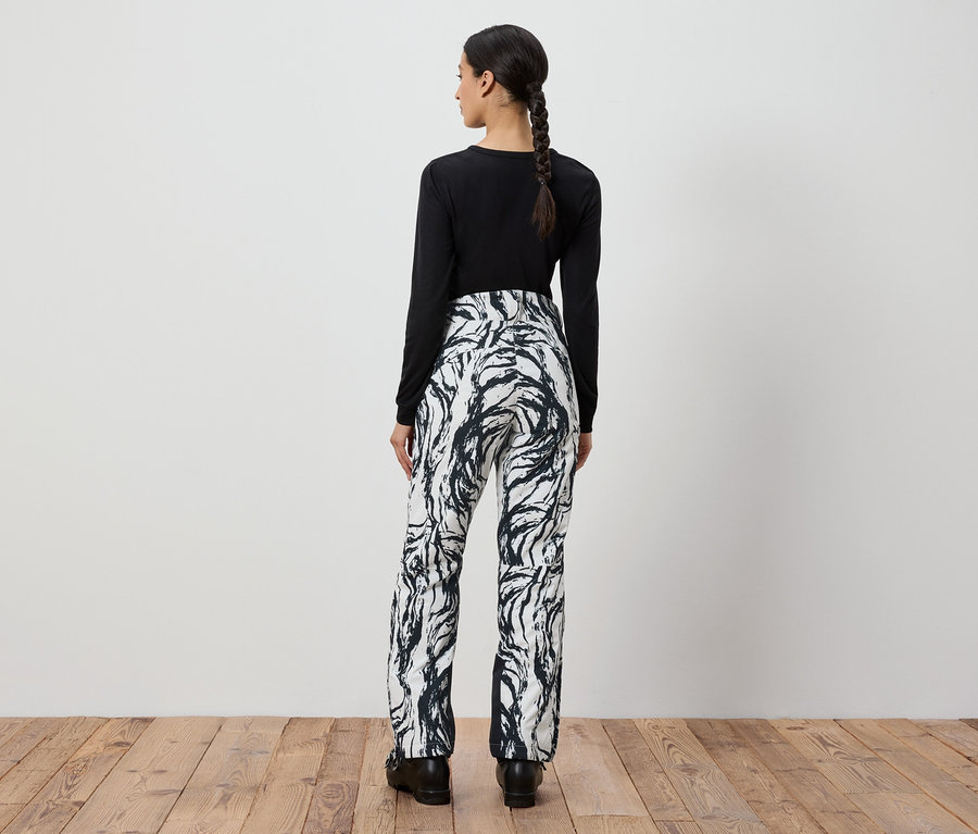 Frau mit Zopf von hinten, trägt ein schwarzes Langarmshirt und eine weiß-schwarze Hose mit Muster.