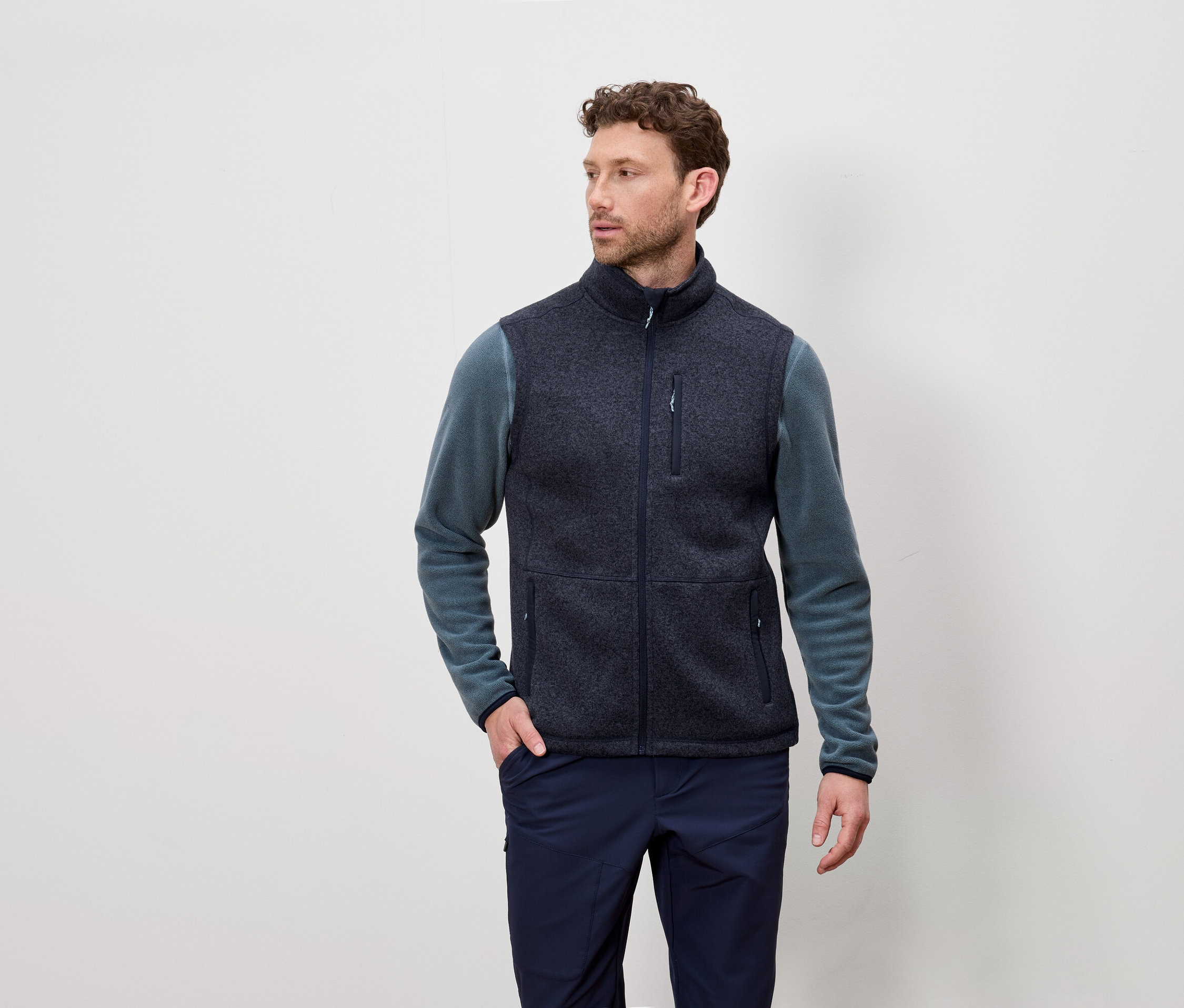 Mann mit kurzen lockigen Haaren trägt eine marineblaue Fleeceweste und ein blaugrünes langärmeliges Hemd und posiert vor einem hellen Hintergrund.