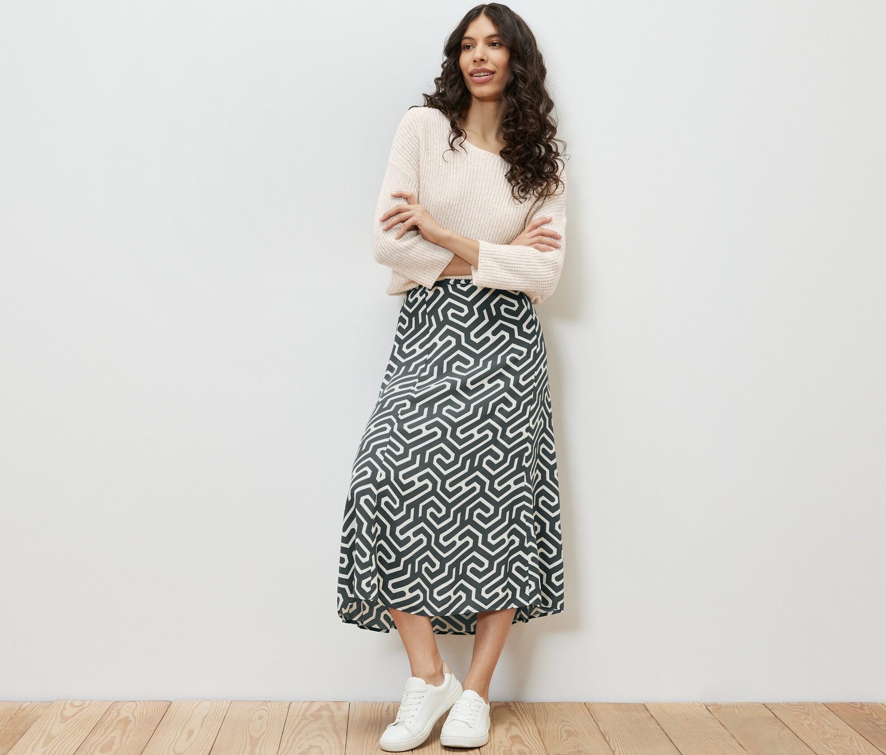 Frau steht mit verschränkten Armen und trägt einen bedruckten Midirock, einen Pullover und Turnschuhe.