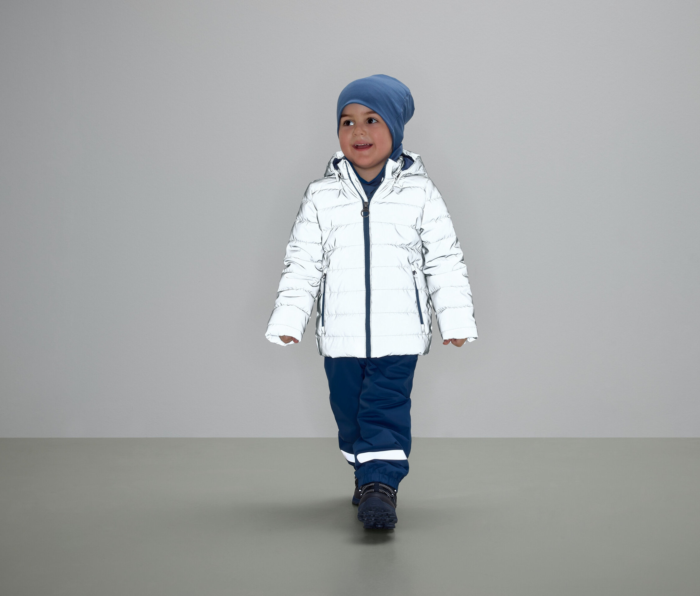 Ein Junge geht in einer weißen Daunenjacke, blauen Hosen und einer Mütze.