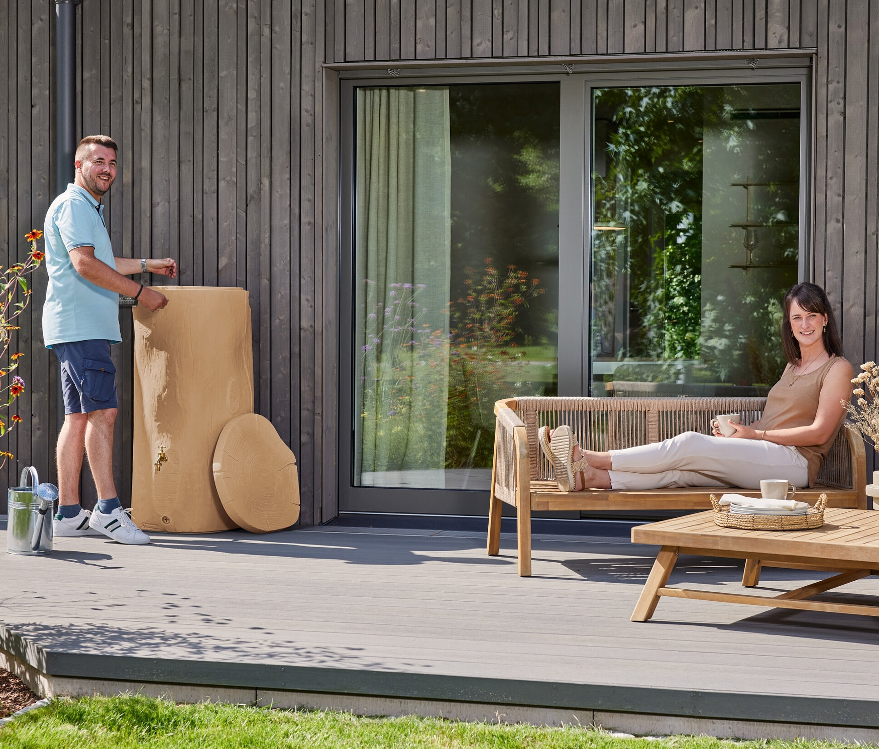 Ein Mann steht neben einem hellbraunen Regenwassertank auf einer Terrasse. Eine Frau entspannt sich auf einer Bank mit einem Getränk.