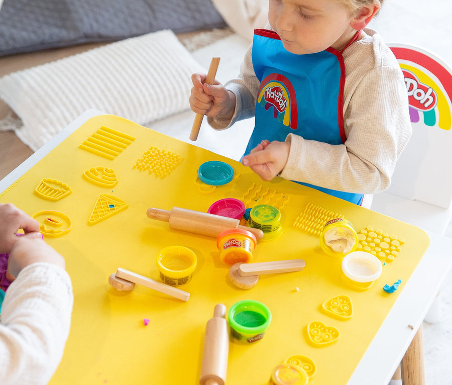 Ein Kind mit Play-Doh-Lätzchen spielt an einem gelben Tisch mit Play-Doh-Knete, Werkzeugen und Förmchen.