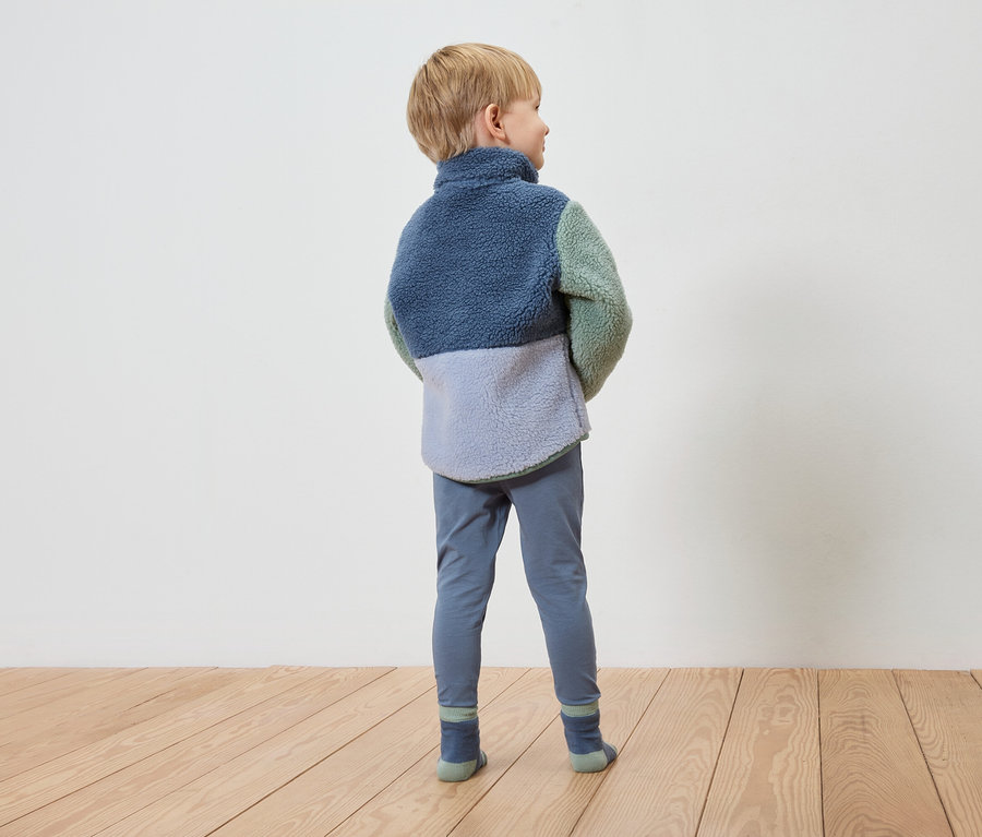 Ein Junge mit blonden Haaren steht mit dem Rücken zur Kamera und trägt eine Fleecejacke in Blau, Grün und Grau, blaue Leggings und Socken.
