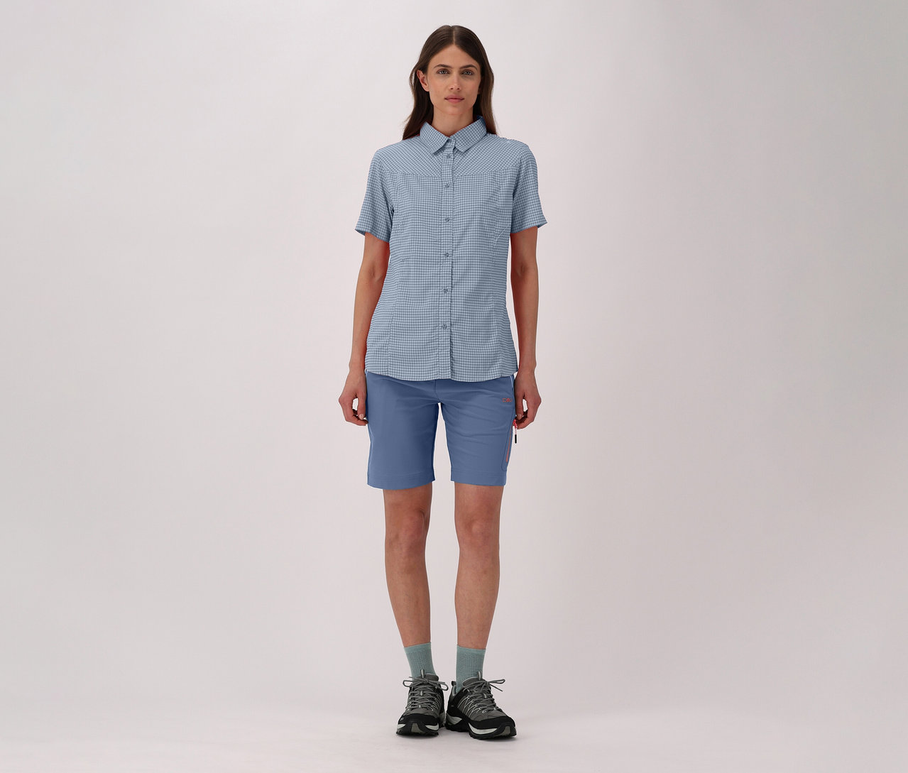 Frau in karierter CMP Damen-Outdoorbluse, blauen Shorts, Socken und Wanderschuhen steht vor hellem Hintergrund.