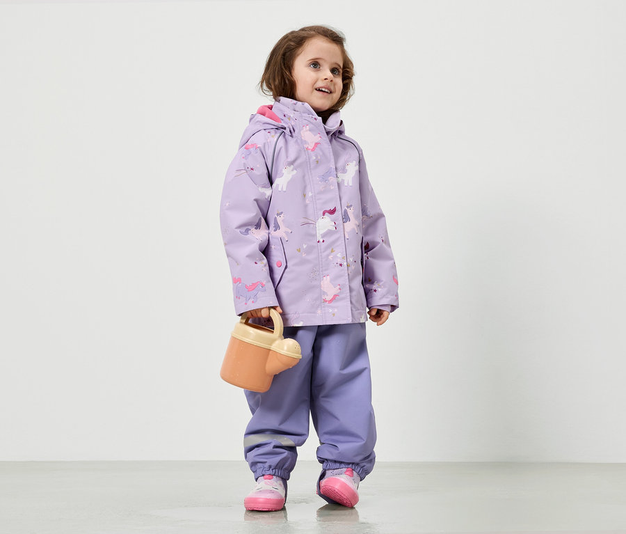 Ein Mädchen in lila Regenkleidung mit Einhornmuster hält eine Gießkanne.
