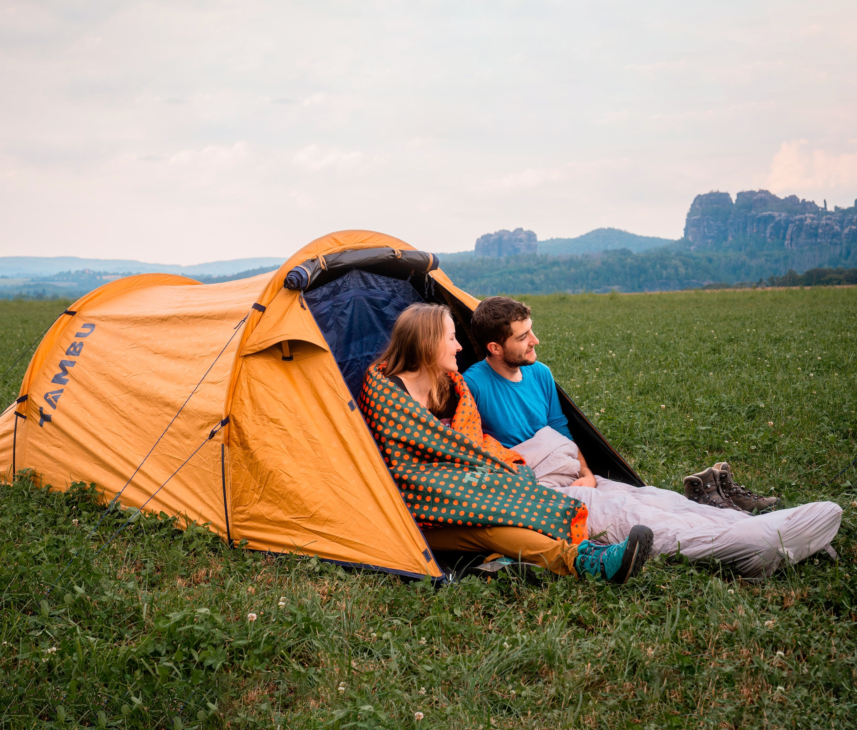 Zwei Personen sitzen in einem orangefarbenen Zelt, wobei eine Person in einen Tambu »KARA 1.600« Mumienschlafsack gehüllt ist.