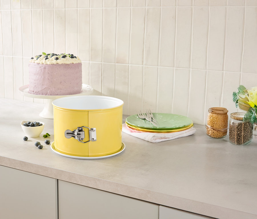 Auf einer Küchentheke stehen eine gelbe Springform, ein Kuchen auf einem Tortenständer, Teller und Vorratsgläser.