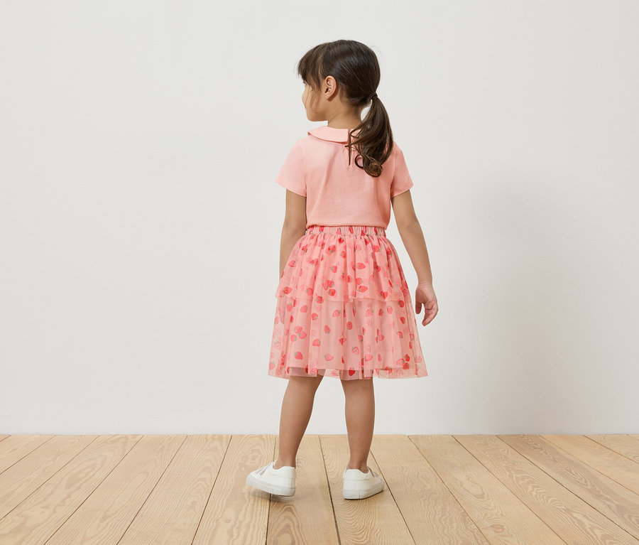 Ein junges Mädchen steht mit dem Rücken zur Kamera, trägt ein rosa T-Shirt, einen rosa Tüllrock mit Erdbeermuster und weiße Turnschuhe.