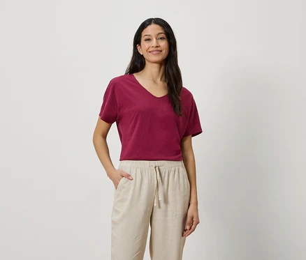 Frau mit dunklen Haaren, die ein weinrotes T-Shirt und eine beigefarbene Hose trägt.