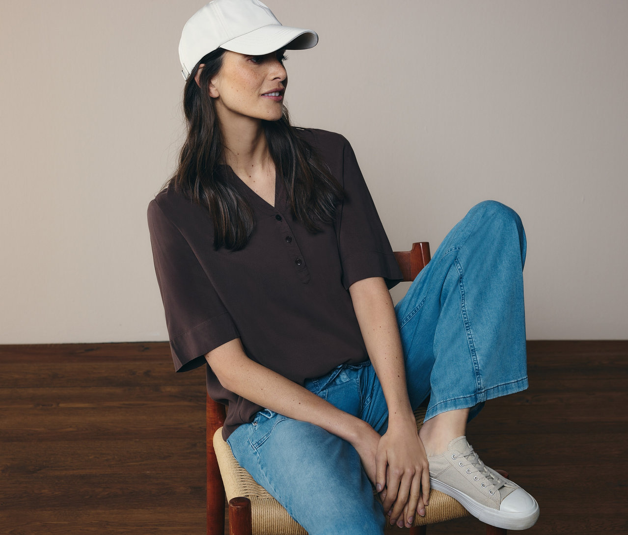 Frau sitzt auf Stuhl, trägt Jeans, Cap und braune CECIL Tunikabluse.