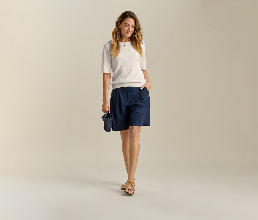 Frau mit Leinen-Bermuda-Shorts, dunkelblau, und Sandalen. Sie hält eine dunkelblaue Mütze in der Hand.