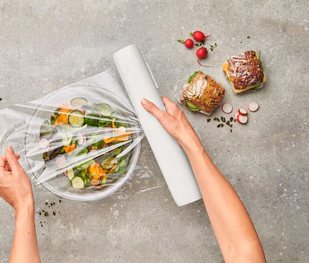 Draufsicht: Person verpackt Salat in Schüssel mit Frischhaltefolie. Daneben: Dressing, belegte Brote und Radieschen.