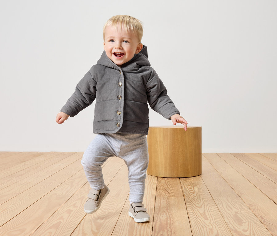 Kleiner Junge mit blonden Haaren, grauer Jacke mit Kapuze, grauer Hose und beigen Schuhen, der auf einem hellen Holzboden läuft.
