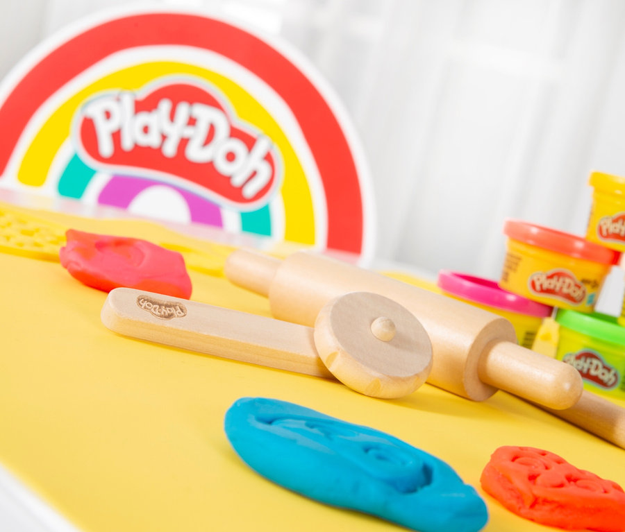Play-Doh-Knete, Holzroller und Formen liegen auf gelbem Untergrund vor einem Play-Doh-Regenbogen.