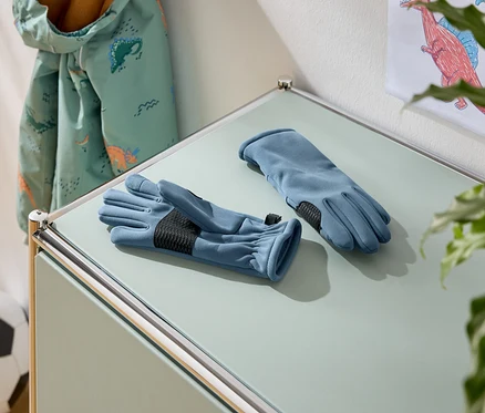 Auf einem Tisch liegen zwei blaue Handschuhe mit schwarzen Handflächen. Im Hintergrund sind eine Jacke mit Dinosaurier-Muster und ein Fußball zu sehen.