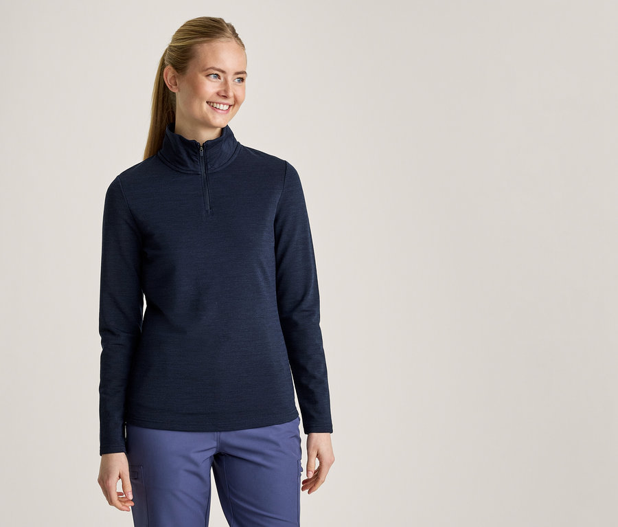 Lächelnde Frau mit Pferdeschwanz, die ein dunkelblaues Langarmshirt mit halbem Reißverschluss und hellblaue Hosen trägt.