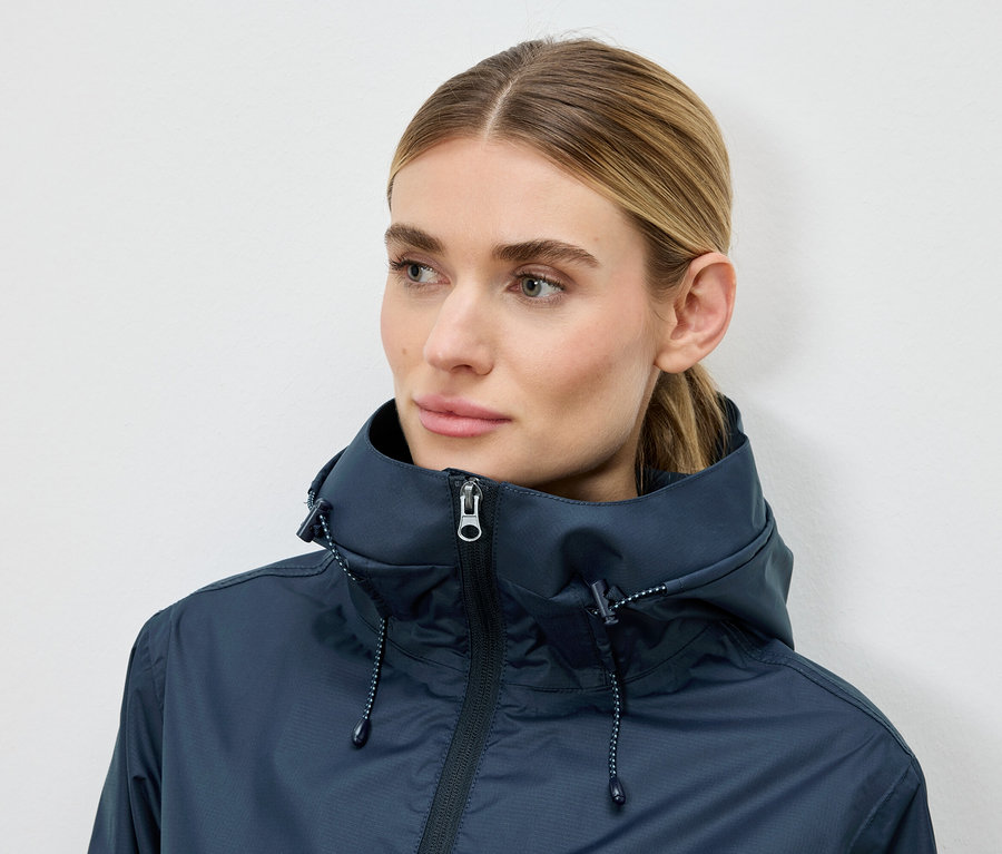 Frau trägt eine dunkelblaue Jacke mit Kapuze vor einem neutralen Hintergrund.