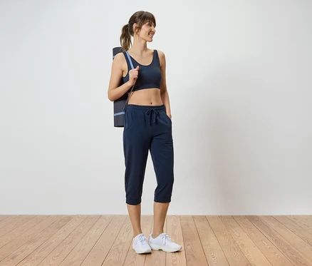 Frau mit dunklem Sport-BH, Dreiviertel-Hose und Yogamatte über der Schulter.