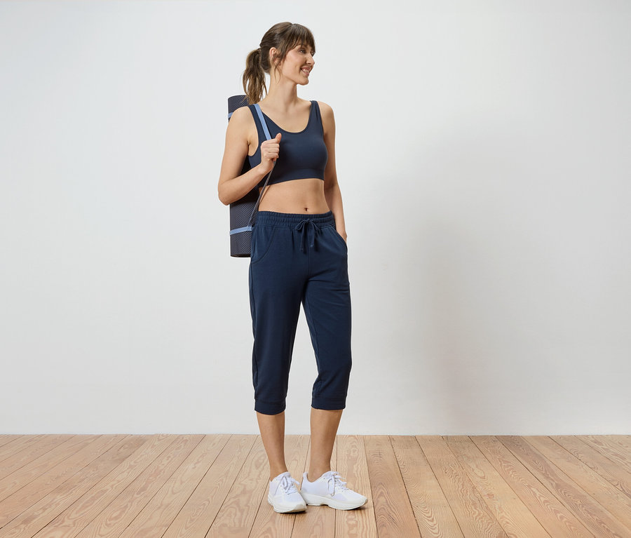 Frau mit dunklem Sport-BH, Dreiviertel-Hose und Yogamatte über der Schulter.